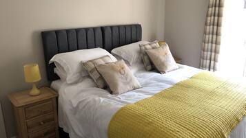 Un lit bien fait, avec du linge de lit blanc et une couverture jaune matelassée, flanqué de deux tables de chevet avec des lampes, dans une pièce dont la tête de lit est sombre et les murs sont de couleur unie.
