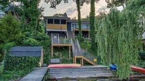 A waterfront property with a wooden dock, a staircase leading to a balcony, and a weeping willow tree.