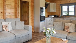 A cozy cabin interior with two grey sofas, a wooden ceiling, a kitchen with a microwave and oven, and a window with a view.
