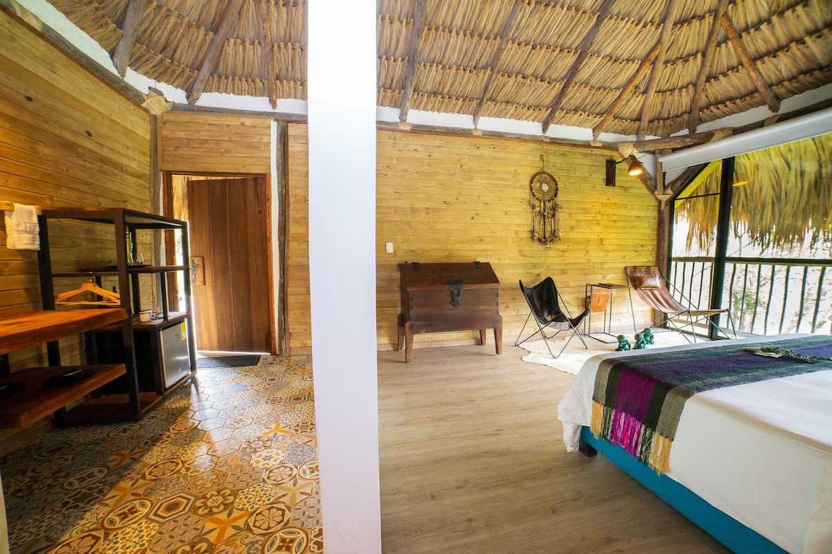 Une pièce avec un plancher en bois, un lit recouvert d’une couverture colorée, un bureau en bois, une chaise et un toit de chaume.