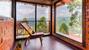 Un balcón con una silla de madera y un florero, con vistas a un paisaje exuberante con árboles y colinas.