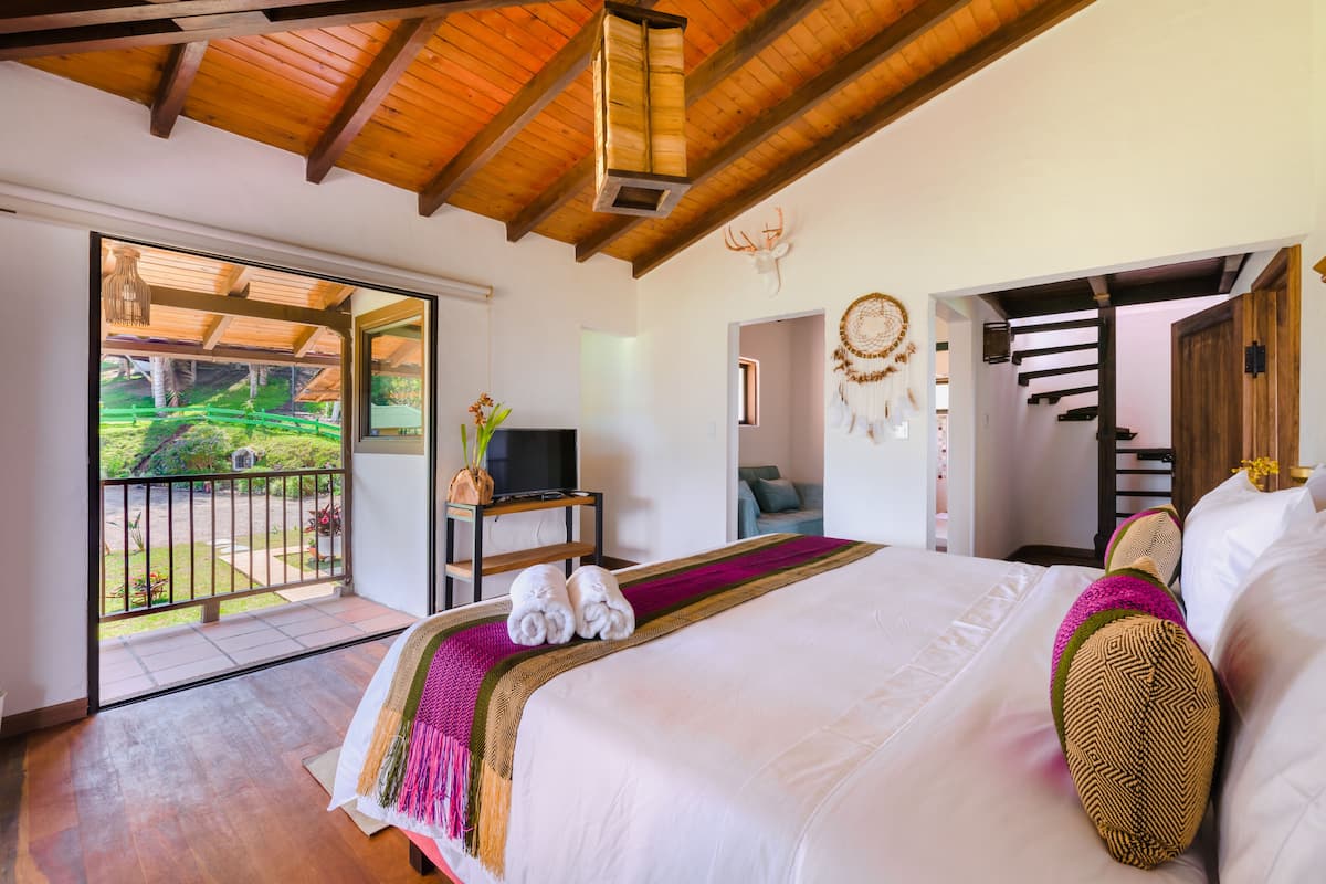 Une chambre spacieuse avec un grand lit, un téléviseur posé sur une table de chevet et un balcon offrant une vue sur la verdure.