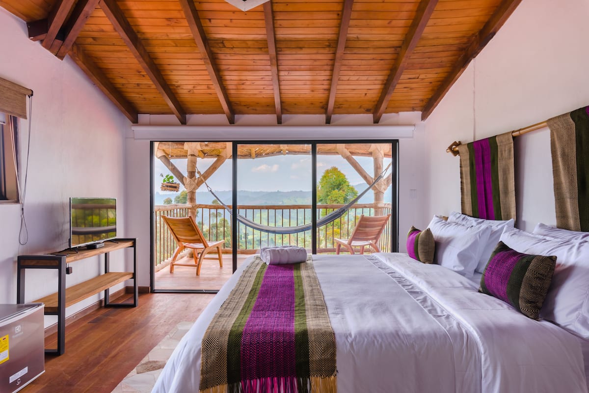 Une chambre avec un grand lit, une télévision, un plafond en bois et un balcon équipé de chaises et d’un hamac.