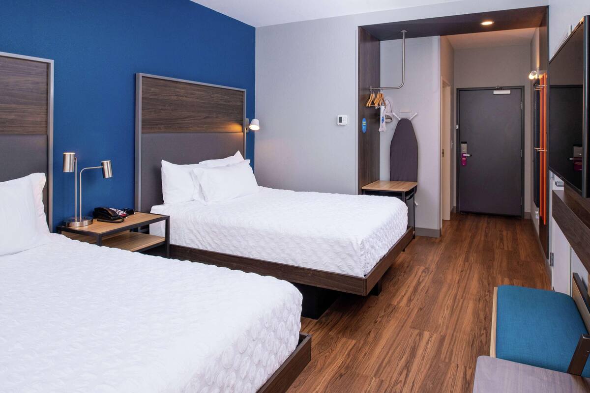 A hotel room with two beds, a wooden floor, a blue accent wall, and a mirror.