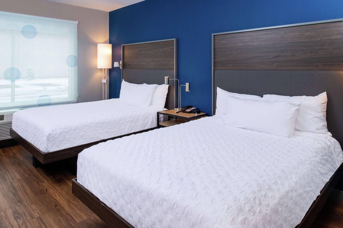 A hotel room with two single beds, a nightstand with a lamp, a telephone, and a window with blinds.