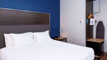 Una cama bien hecha con ropa blanca, un cabecero de madera, una mesita de noche con una lámpara, y un perchero con una toalla y una prenda colgada.