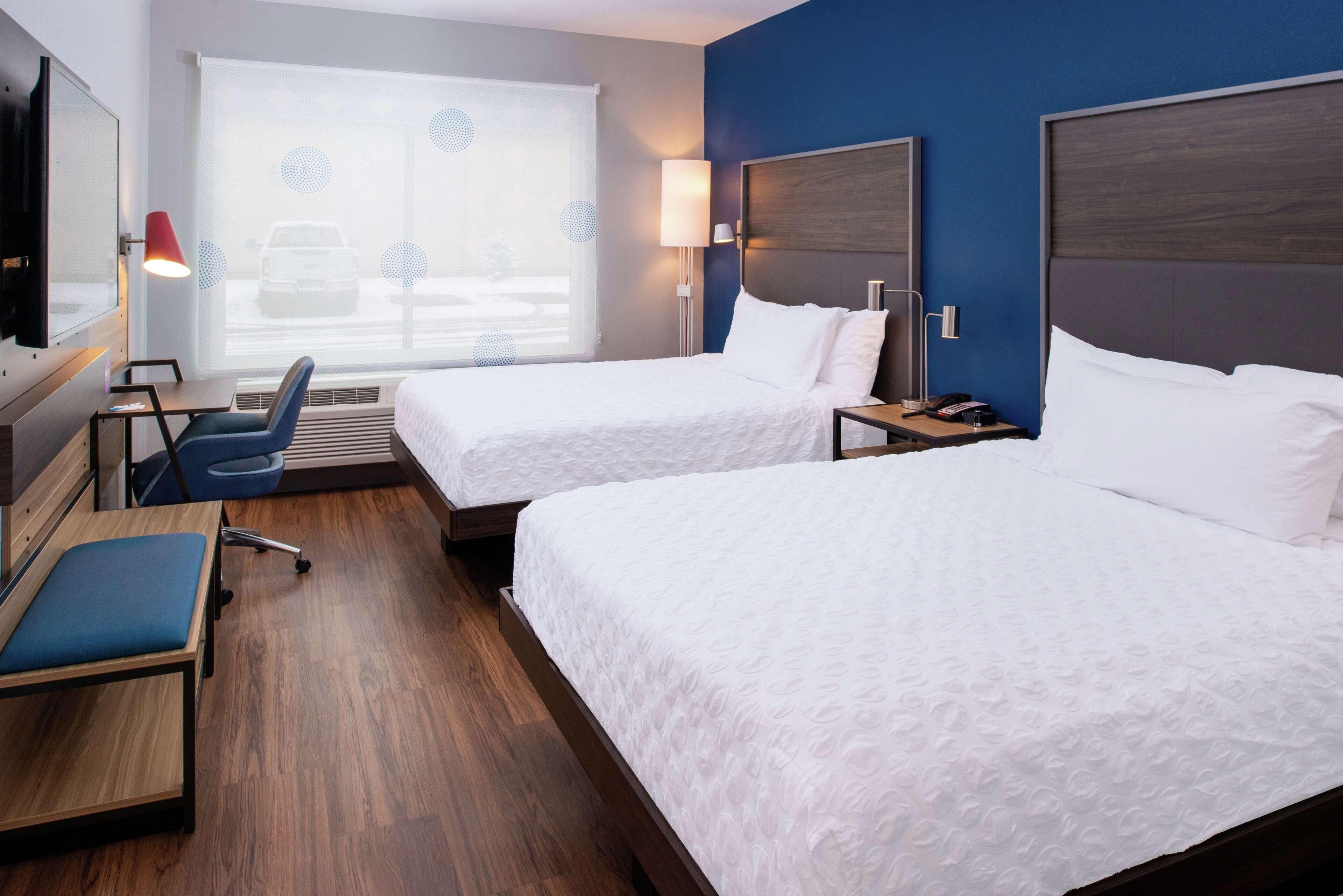 A hotel room with two beds, a desk, a chair, and a large window with blinds.