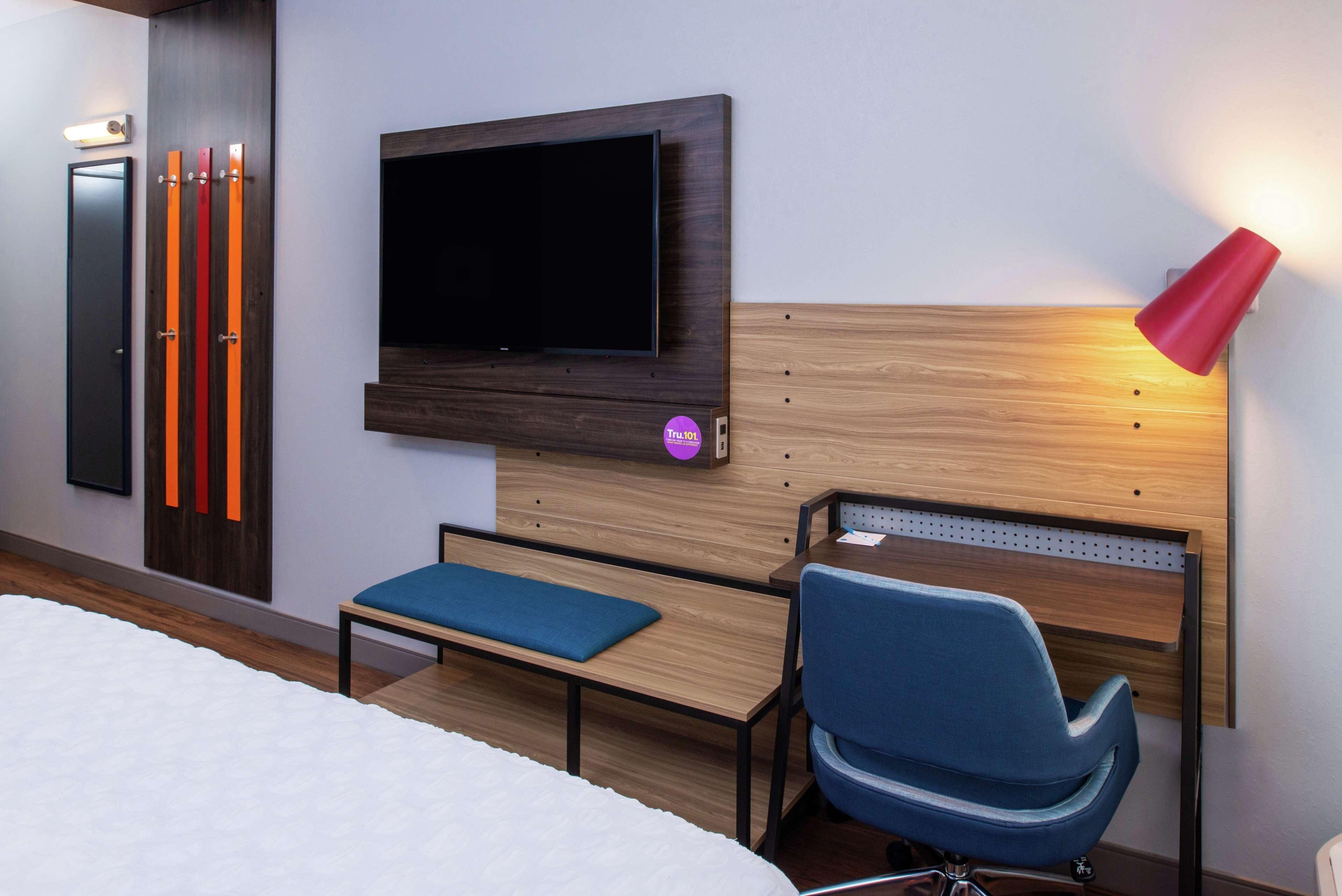 A hotel room with a bed, a TV mounted on the wall, a blue chair, a wooden headboard, and a red lamp.