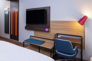 A hotel room with a bed, a TV mounted on the wall, a blue chair, a wooden headboard, and a red lamp. - Tru By Hilton Elkhart (Elkhart)