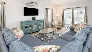 A living room with a grey sectional sofa, a round coffee table, a teal cabinet, a TV mounted on the wall, and a window with curtains. - 5 Bedroom Oceanview House with a Private Pool! Stingray (Surfside Beach)