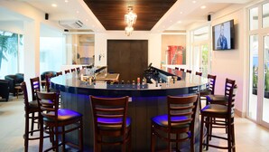 A modern bar area with a central bar, multiple seating options, and a large screen on the wall. - Gran Costa Azul Hotel (Puerto Barrios)