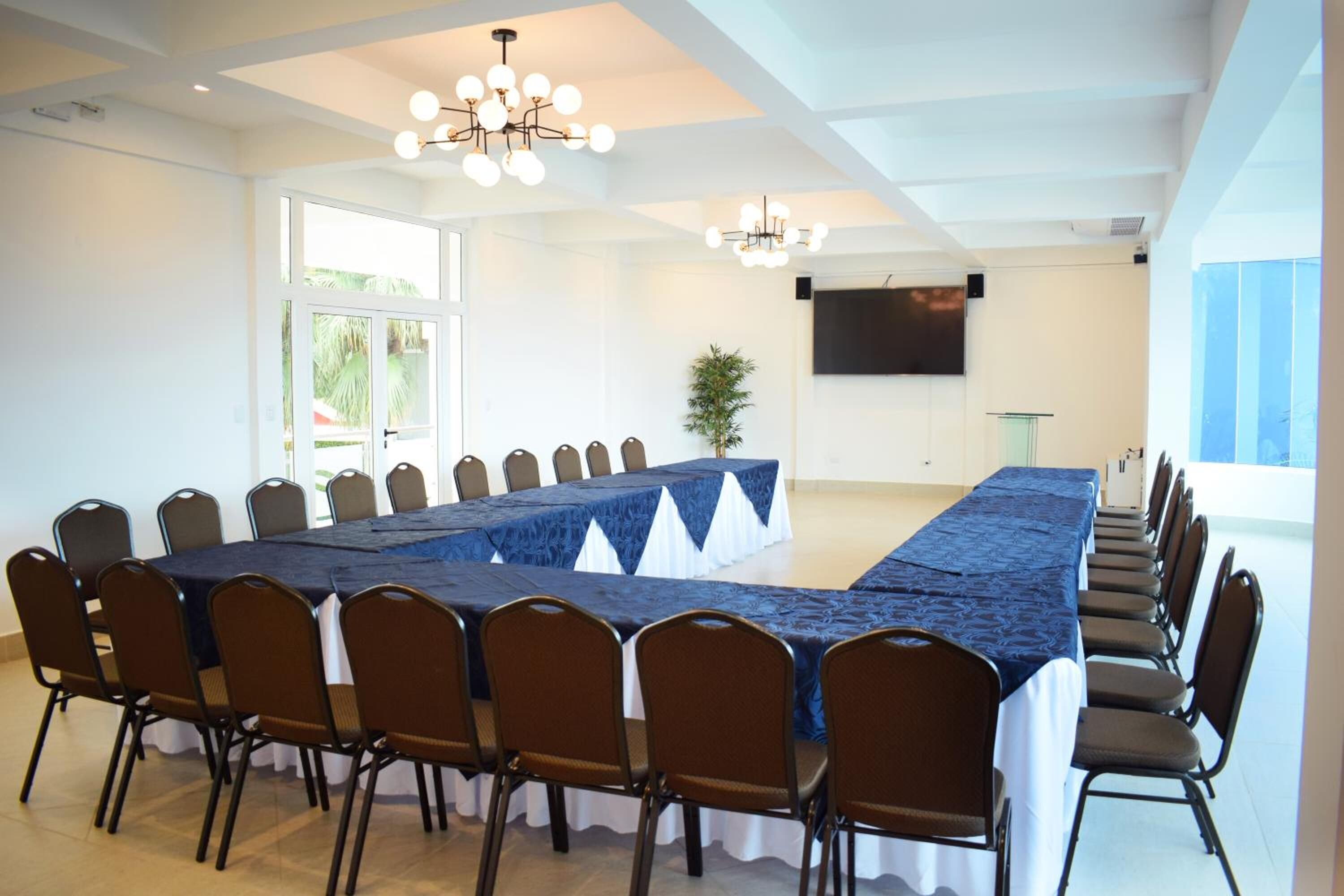 Un salón de conferencias con una larga mesa, sillas, candelabros, una pantalla de televisión y un ventanal.