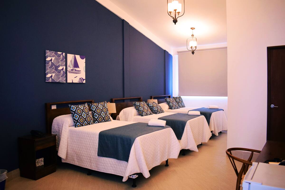A hotel room with three beds, each with a blue headboard and white bedding. There are two wall-mounted lamps and a ceiling light.