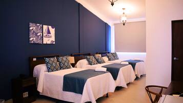 Habitación de hotel con tres camas, cada una con cabecero azul y ropa de cama blanca. Hay dos lámparas de pared y una lámpara de techo.