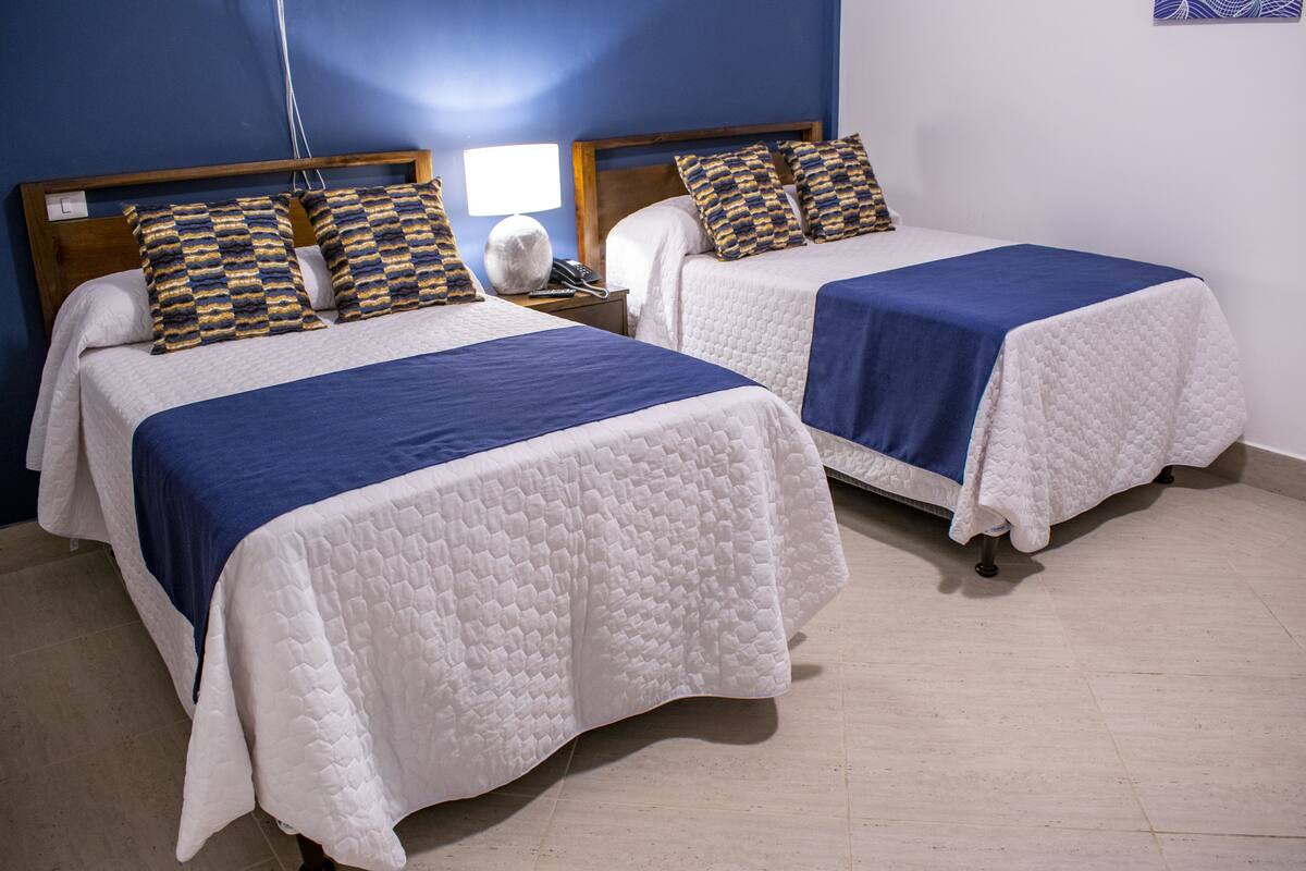 Two single beds with blue and white bedding, a bedside table with a lamp, and a wall socket.