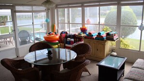 Ein Wintergarten mit Glastisch, Korbstühlen und Blick auf einen See, sowie eine Auswahl an Spielzeug und Büchern für Kinder auf Regalen.