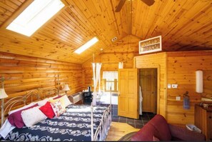 Cozy cabin-style room with a high ceiling and wooden beams, featuring a bed with a floral-patterned bedspread and a small table with a lamp.