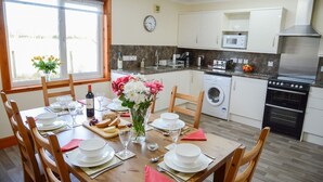 A dining area with a wooden table set for a meal, a washing machine, a microwave, and a stove in the kitchen. - Tigh Grianach -  a cottage that sleeps 6 guests  in 3 bedrooms (Oban)