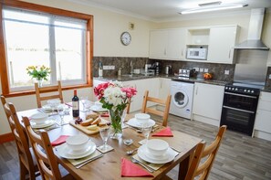 A dining area with a wooden table set for a meal, a washing machine, a microwave, and a stove in the kitchen. - Tigh Grianach -  a cottage that sleeps 6 guests  in 3 bedrooms (Oban)