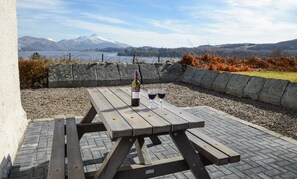 An outdoor picnic table with a bottle and glasses, set against a backdrop of mountains and a lake. - Tigh Grianach -  a cottage that sleeps 6 guests  in 3 bedrooms (Oban)