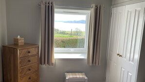 A bedroom with a bed, a wooden dresser, a small stool, and a window with curtains. - Tigh Grianach -  a cottage that sleeps 6 guests  in 3 bedrooms (Oban)