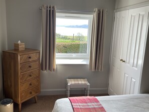 Une chambre à coucher avec un lit, une commode en bois, un petit tabouret et une fenêtre avec des rideaux.