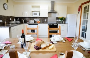 A dining table set with a loaf of bread, cheese, grapes, and a bottle of wine, with a kitchen in the background.