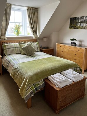 A bedroom with a bed, a wooden dresser, a nightstand, a lamp, a window with curtains, and a painting on the wall.