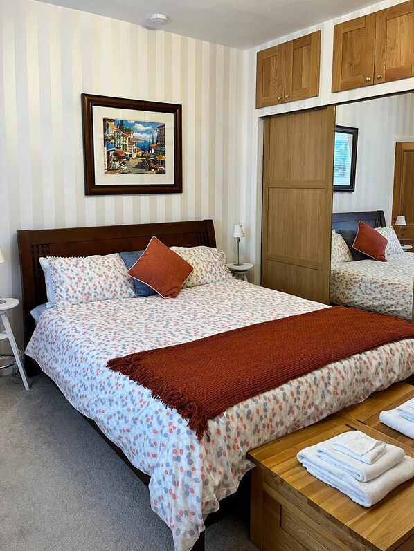 A hotel room with a large bed, a wooden headboard, a bedside table with towels, a nightstand, a framed picture on the wall, and a wardrobe with a mirror.