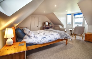 A bedroom with a sloped ceiling, a bed with a patterned duvet, a bedside table with a lamp, a chair, and a view of the outdoors through a window.