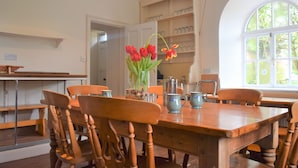 Un comedor con una mesa de madera, sillas y un jarrón con tulipanes rojos. Se observa una estantería con cristalería y una ventana que ofrece vistas a zonas verdes.