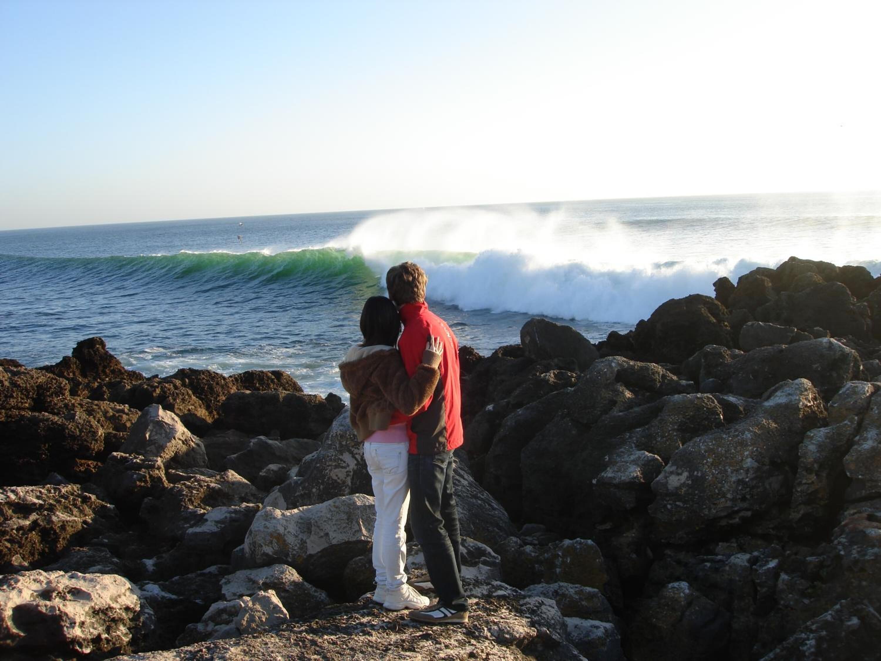 A couple standing on rocky terrain by the ocean, with waves crashing nearby.