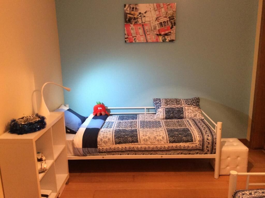Une petite chambre à coucher accueillante, avec un lit simple, une table de chevet, une étagère ornée d’objets décoratifs et un tableau accroché au mur.