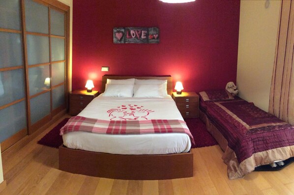 Un dormitorio con una cama grande, dos mesitas de noche con lámparas, una mesita de noche más pequeña con un objeto decorativo, una pared roja y una ventana con cristal satinado.