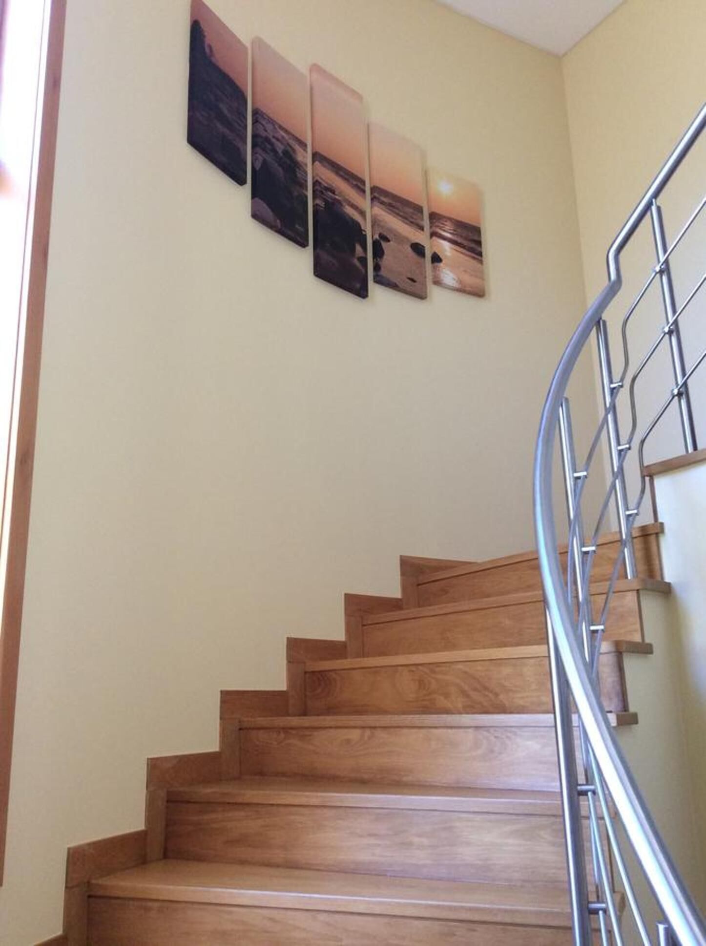 A wooden staircase with a metal handrail, leading to a landing where three framed artworks are displayed on the wall.