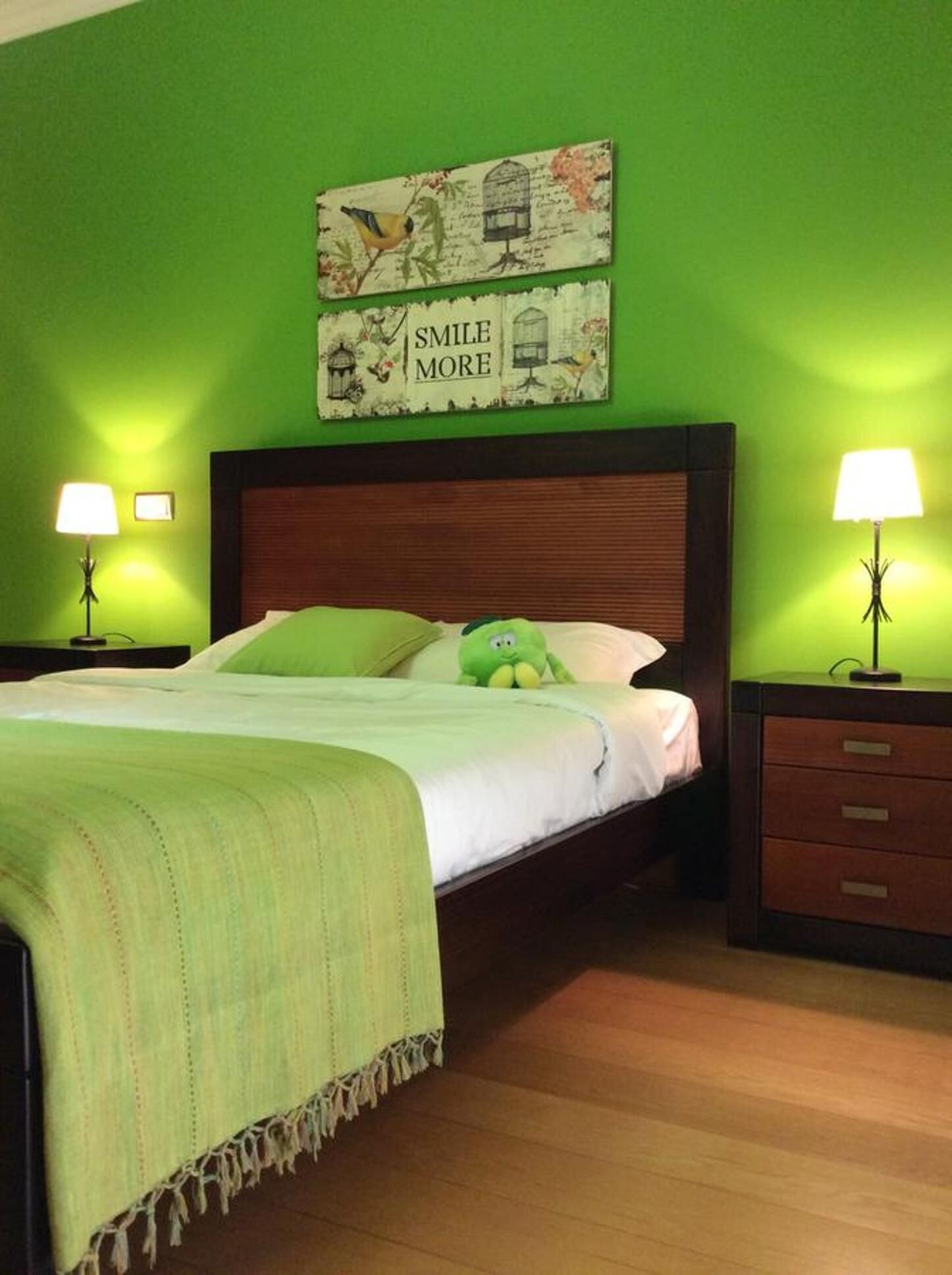 A neatly made bed with a green blanket and a plush toy, two bedside lamps, and a wall with a 'SMILE MORE' sign.