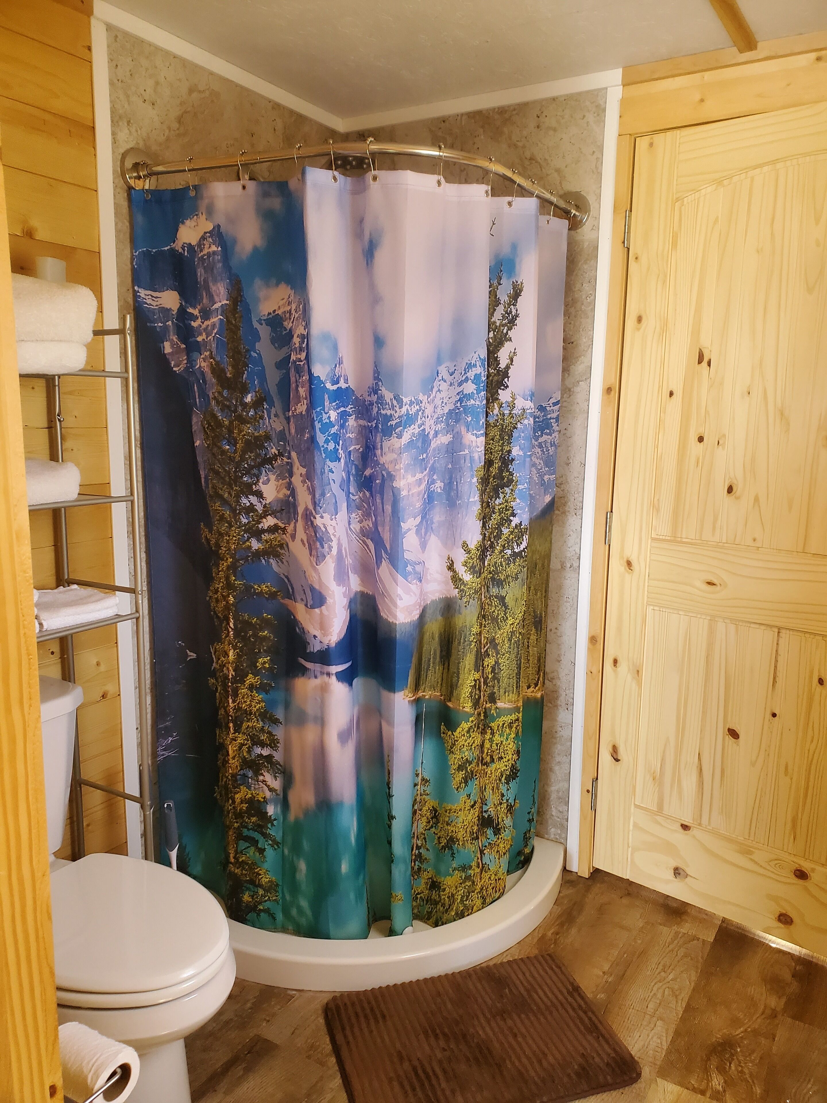 A bathroom with a shower curtain depicting a mountainous landscape, a toilet, and wooden shelving with towels.