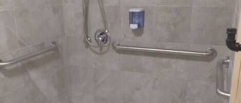 A shower area with a white floor, a handheld showerhead, a wall-mounted faucet, and a handrail.