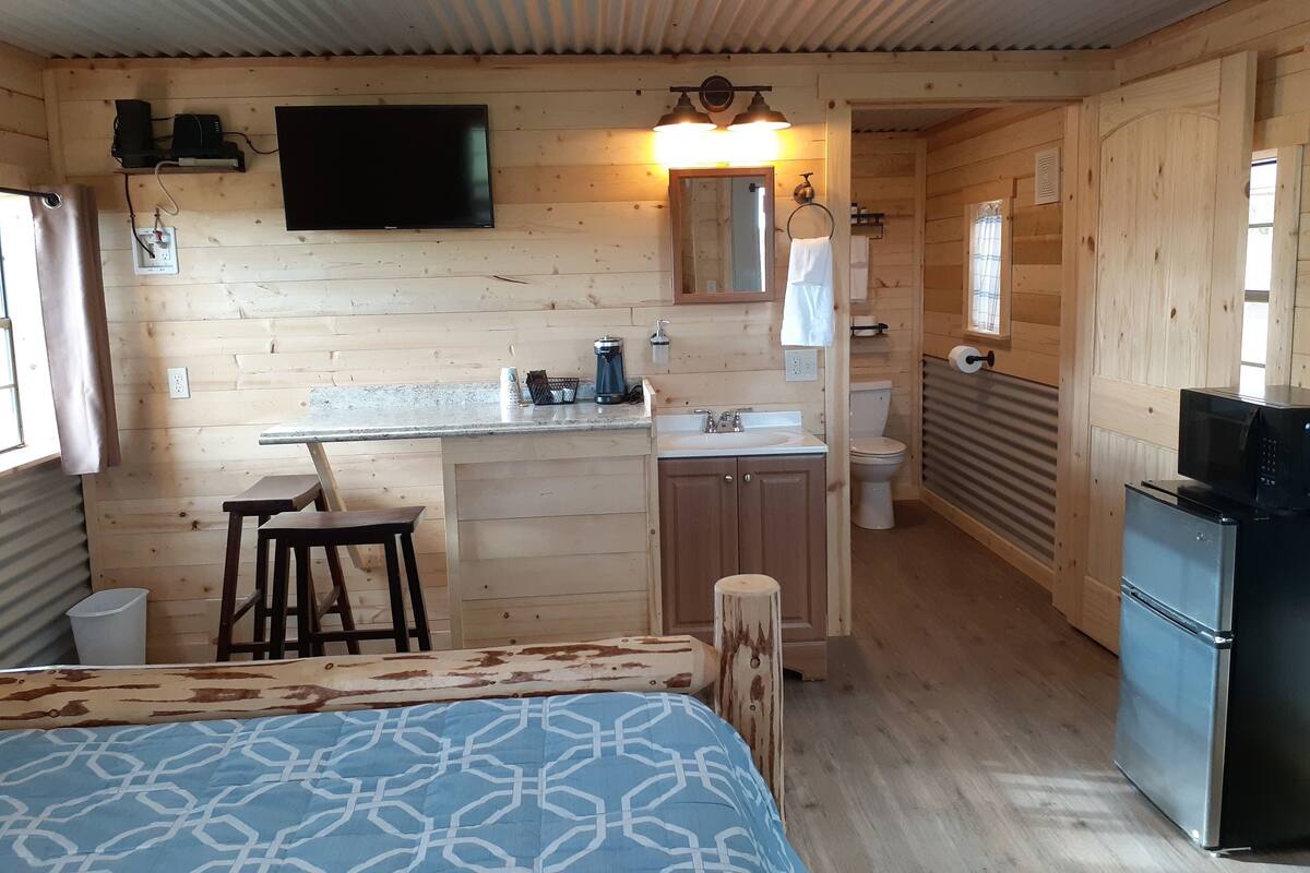 A compact, wood-paneled room with a kitchenette, a bed with patterned bedding, and a television mounted on the wall.