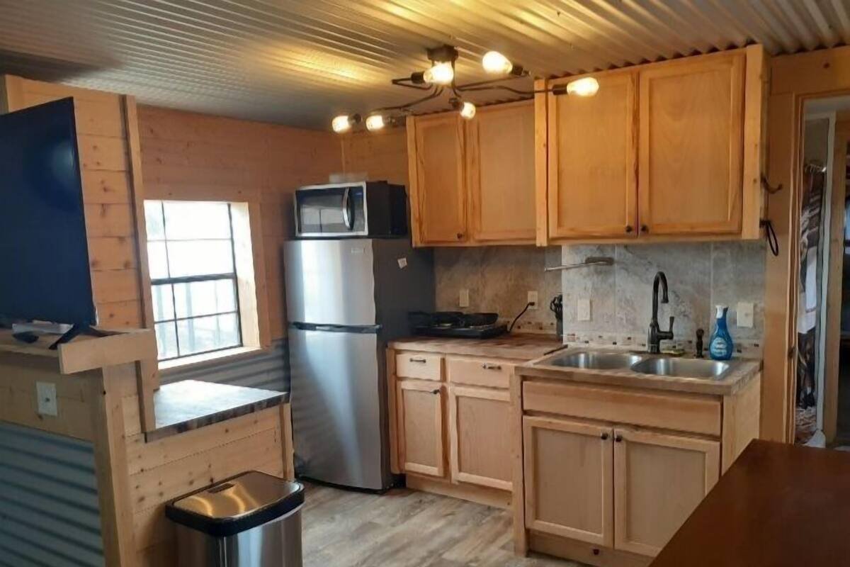 A compact kitchen with wooden cabinets, a stainless steel refrigerator, a microwave, a sink, and a trash bin.
