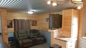 A cozy room with a wooden ceiling, a reclining sofa, a television mounted on a wooden cabinet, and a trash bin. - Glacier Acres Guest Ranch (Columbia Falls)
