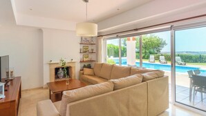 Ein modernes Wohnzimmer mit einem beigefarbenen Ecksofa, einem Holztisch und einem Kamin. Eine große Glasschiebetür führt zu einem Außenpoolbereich.