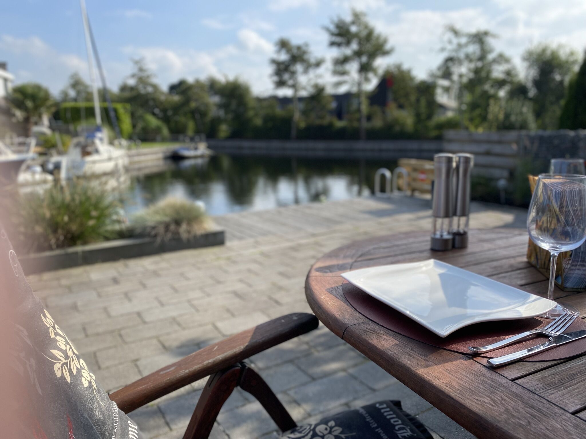 A wooden table with a white plate, a glass of water, and cutlery, set on a patio with a view of a calm water body and boats in the background.