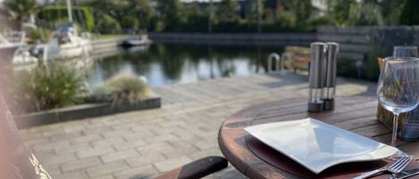 A wooden table with a white plate, a glass of water, and cutlery, set on a patio with a view of a calm water body and boats in the background.