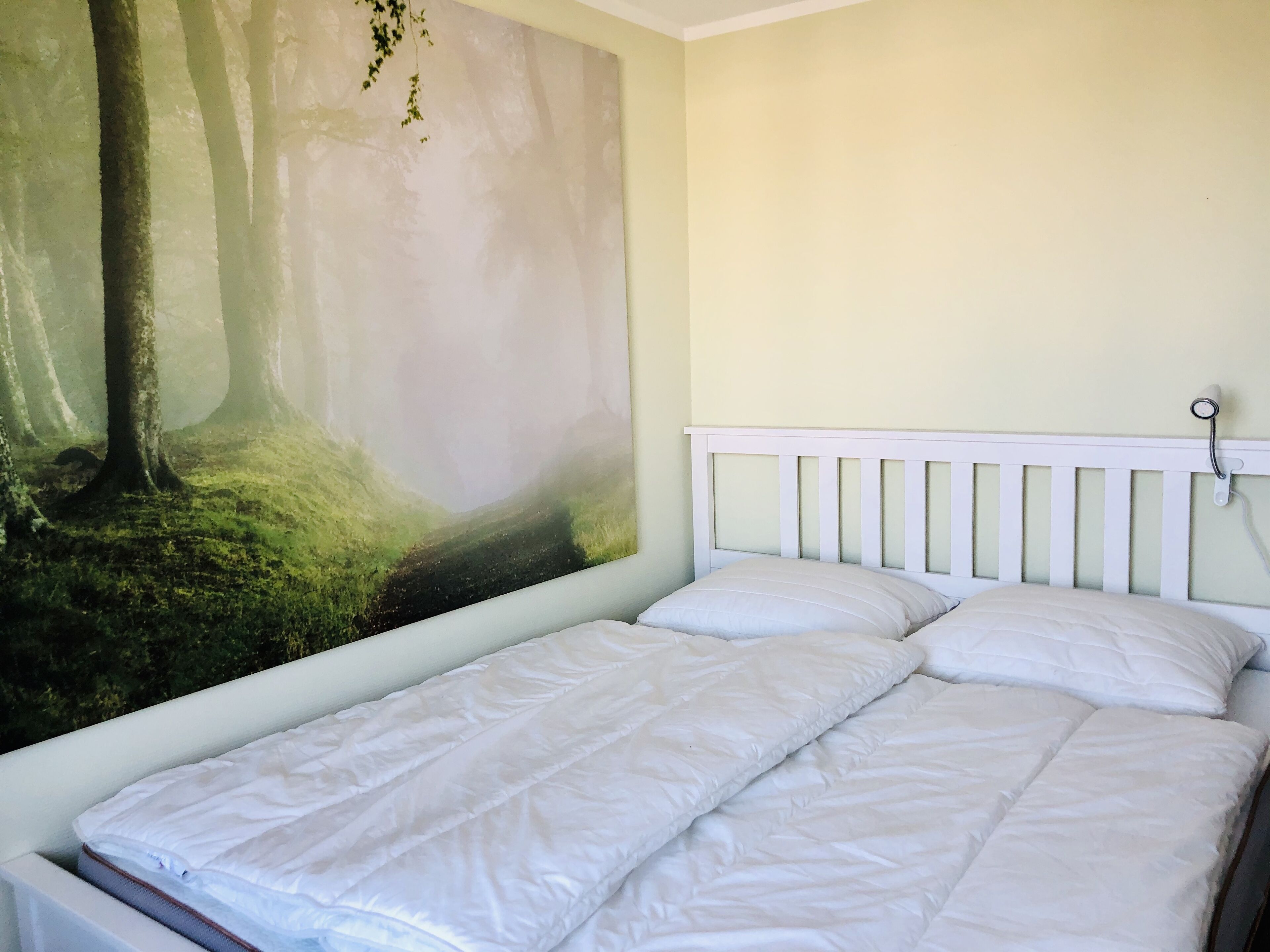 A bedroom with a bed, white bedding, a wall-mounted light, and a forest-themed mural.