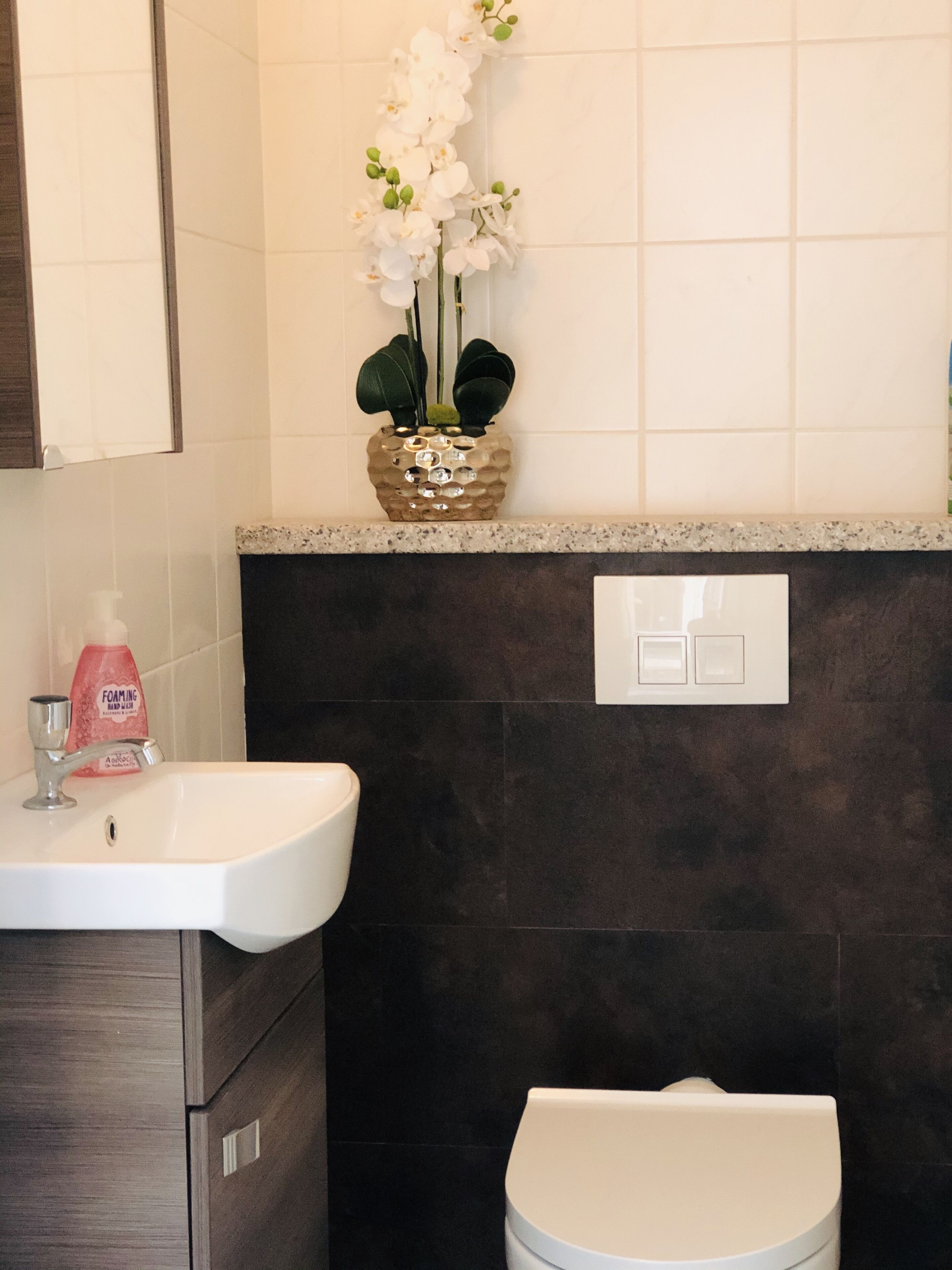 Een badkamer met een witte wasbak, donkere betegelde wanden, een toilet en een decoratieve orchideeënplant in een gepot textuur.