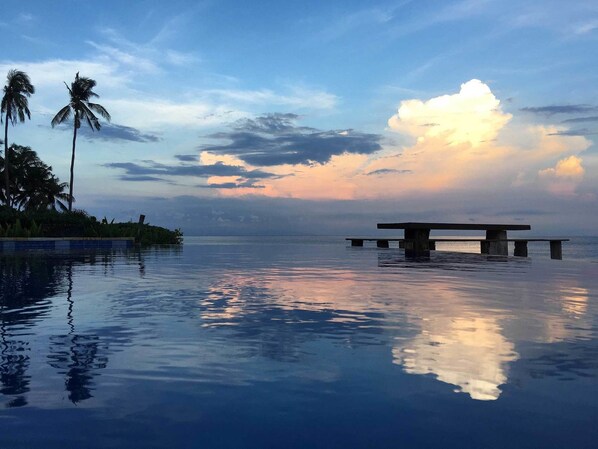 An infinity pool with a wooden bench, palm trees, and a sunset sky. - Jasri Bay Hideaway (Karangasem)