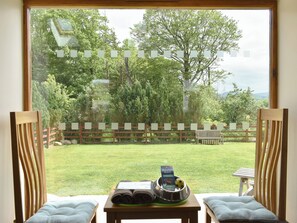 A room with a large window overlooking a grassy yard with trees and a wooden fence. There is a small table with a dog bowl and a rolled-up towel on it. - 2 bedroom accommodation in Ullock, near Cockermouth (Ullock, near Cockermouth)