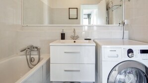 A modern bathroom with a bathtub, a sink with a mirror, and a front-loading washing machine. - Appartement Panorama - Welkeys (Villeneuve-Loubet)