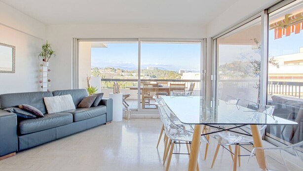 A modern living room with a glass dining table, a grey sofa, and a large window offering a view of the cityscape. - Appartement Panorama - Welkeys (Villeneuve-Loubet)